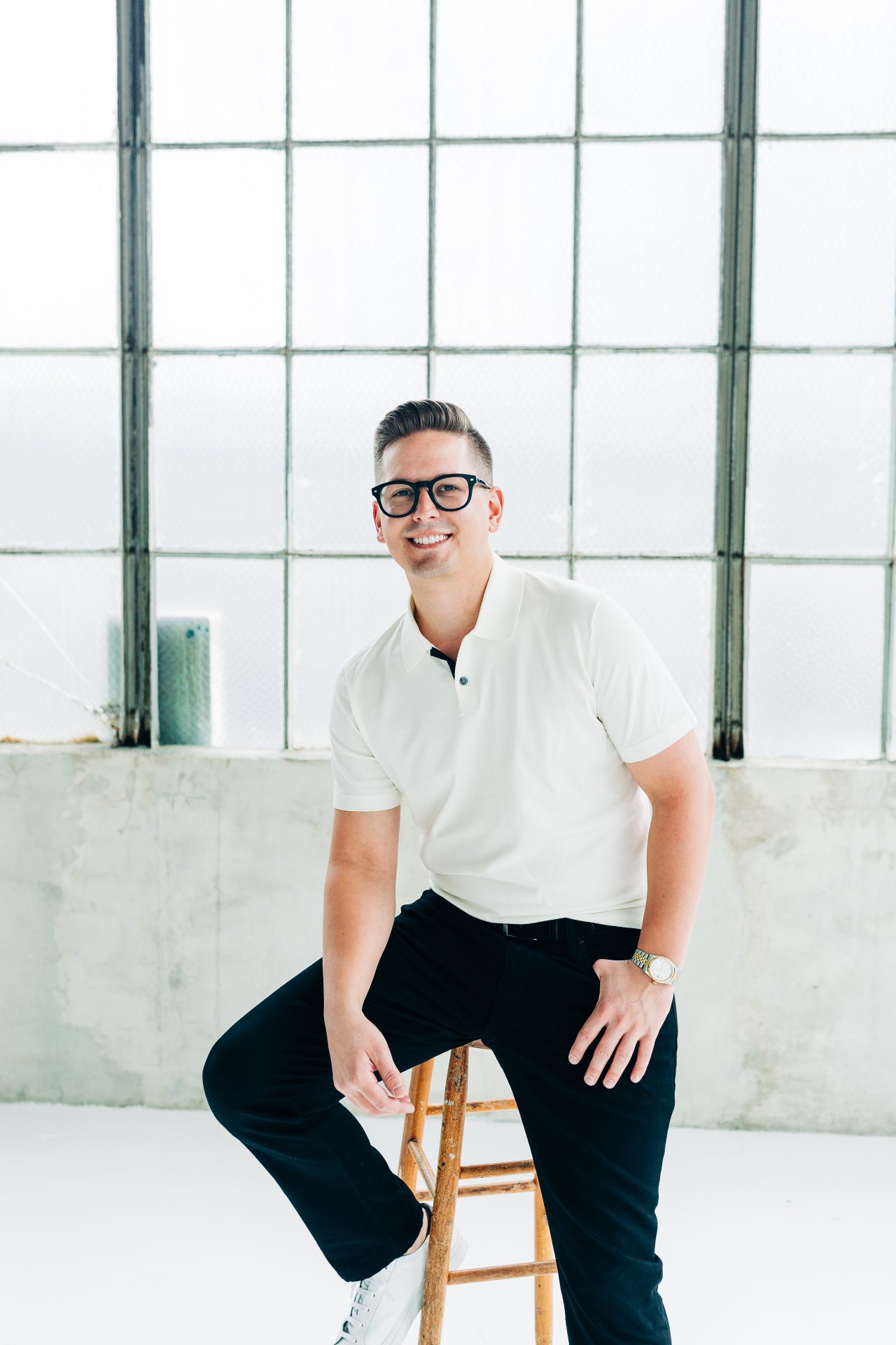 A person wearing glasses and casual clothes sits on a wooden stool in front of large industrial windows. The setting is minimalistic.