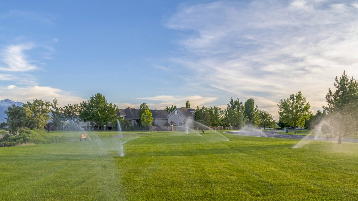 Sprinklers spraying water on green grasses with homes mountain and blue sky view. Picturesque nature and residential scenery with automatic irrigation system.