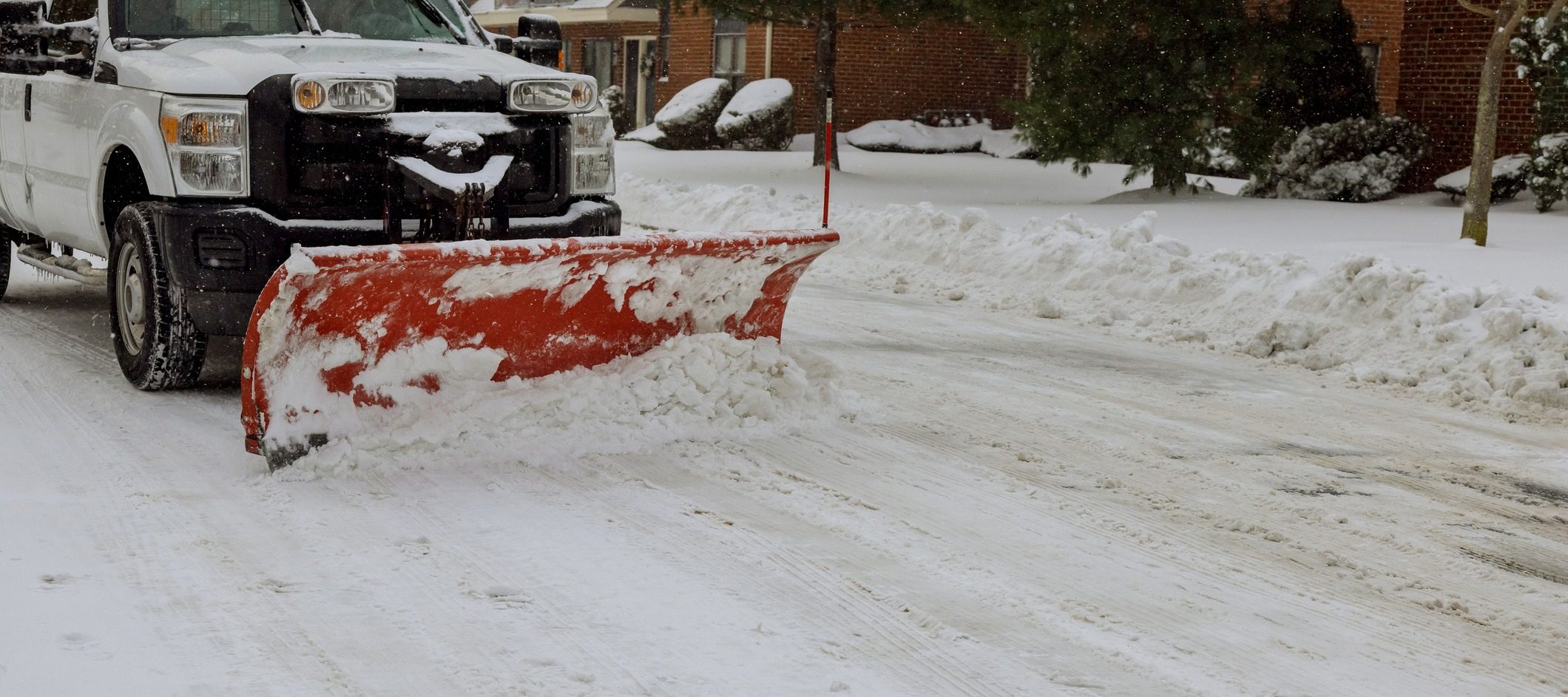 A snow plow clears a snowy residential street, with snow-covered trees and houses visible in the background.