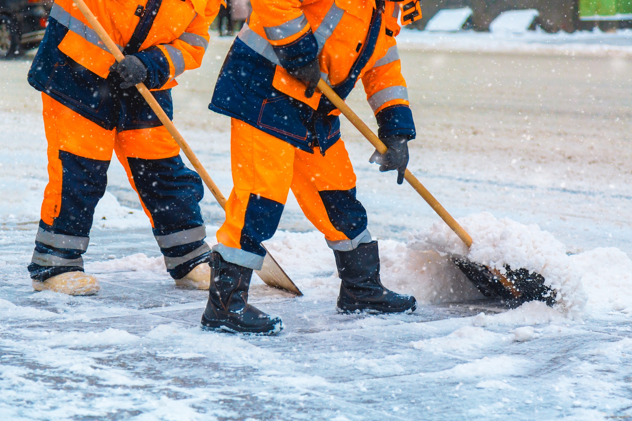 Two people in bright orange gear shovel snow on a snowy street, focusing on clearing the path amid falling snowflakes.