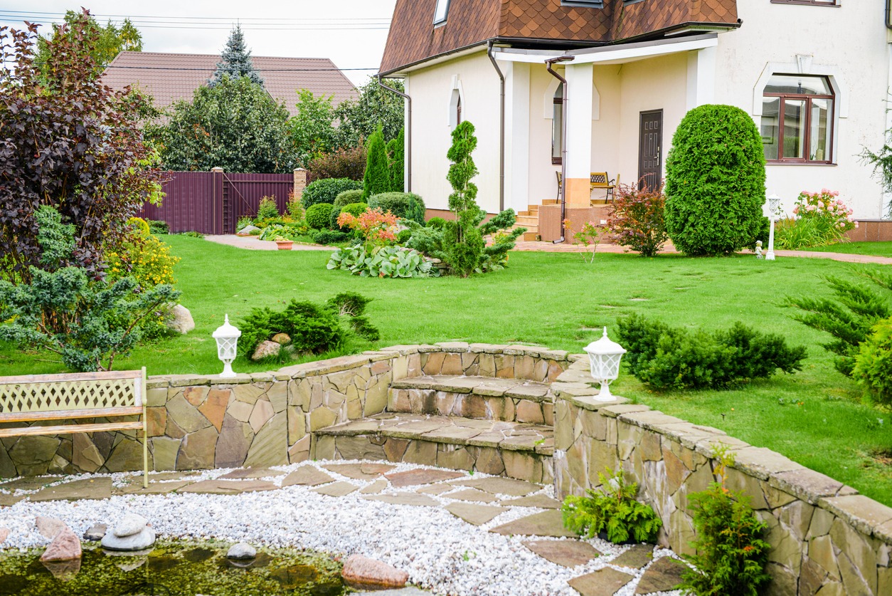 A well-maintained suburban garden with lush greenery, stone path, decorative lanterns, and a modern house in the background, surrounded by trees.