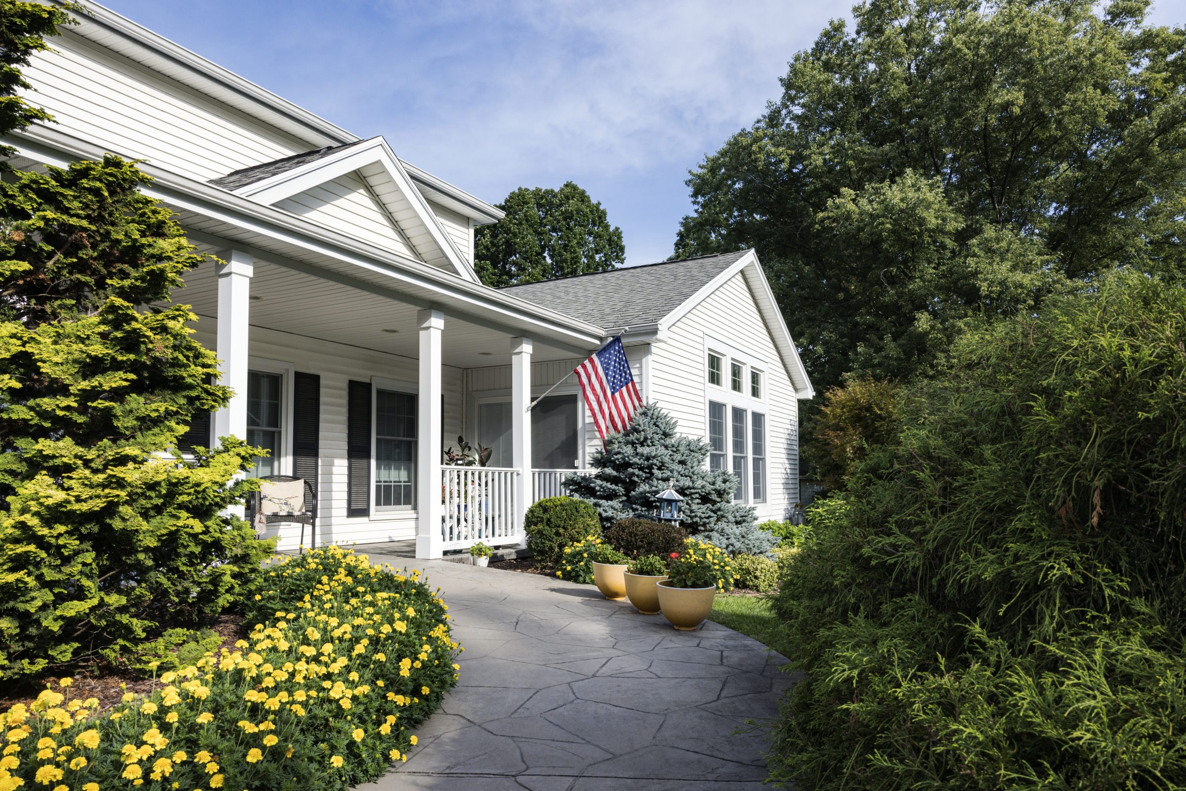 A white house with a porch, American flag, and vibrant garden featuring yellow flowers and potted plants. Surrounded by lush green trees.
