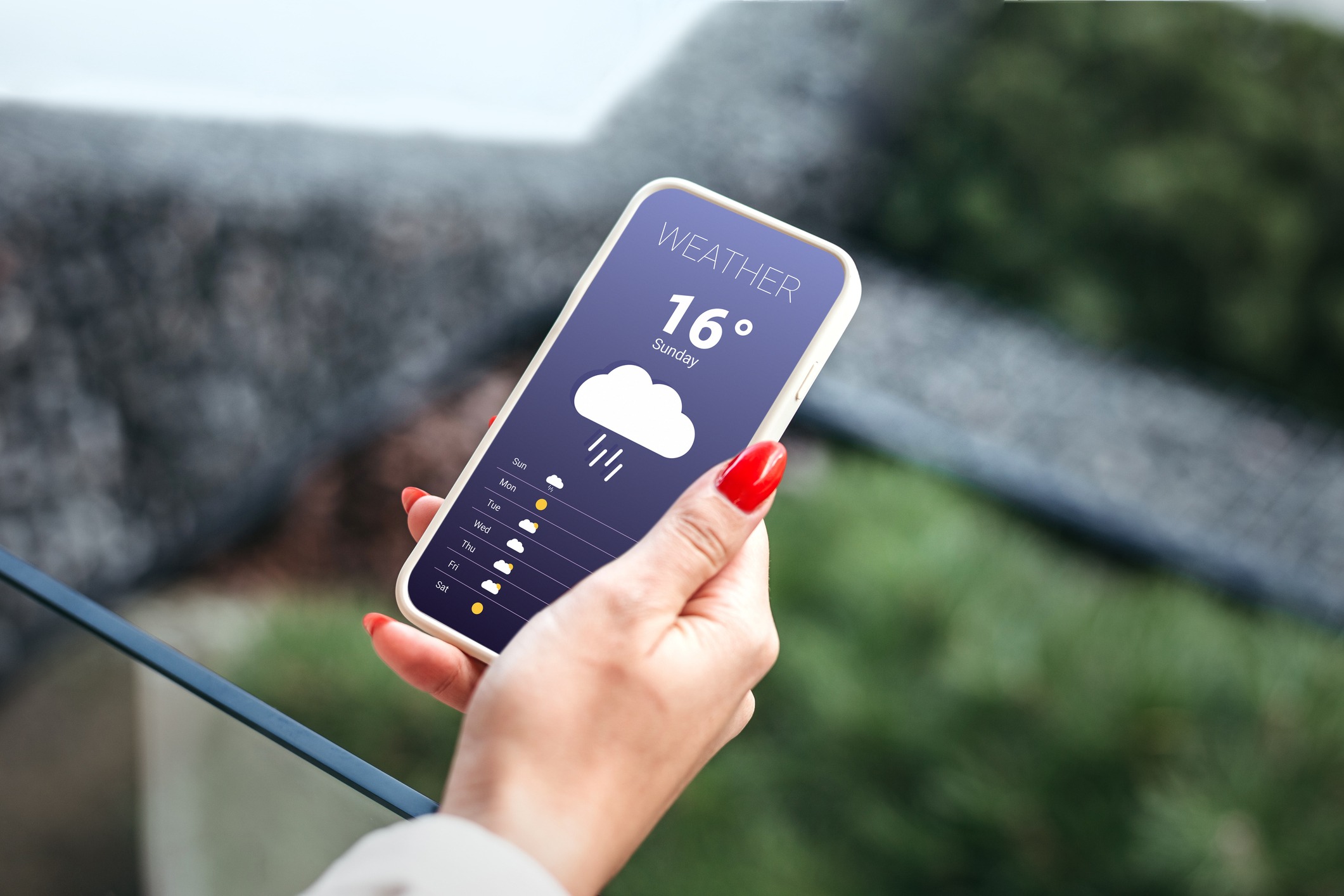 A person holds a smartphone displaying a weather app, showing 16°C with rain. The background is blurred greenery and pavement.