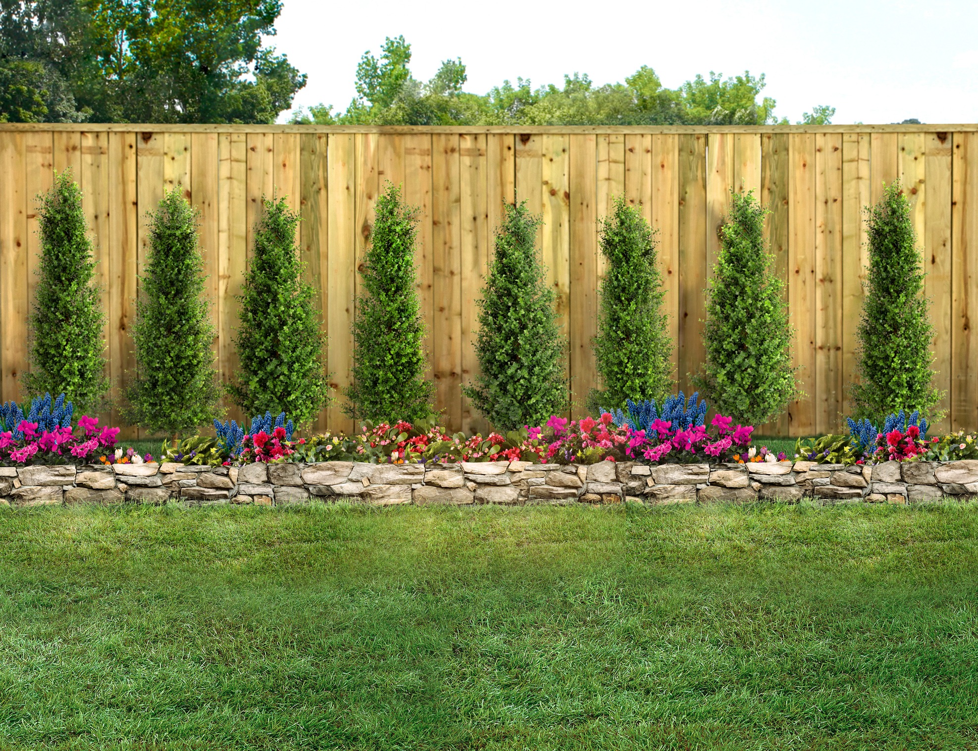 A wooden fence with vertical trees stands behind a colorful flowerbed, outlined by a low stone wall, surrounded by lush green grass.