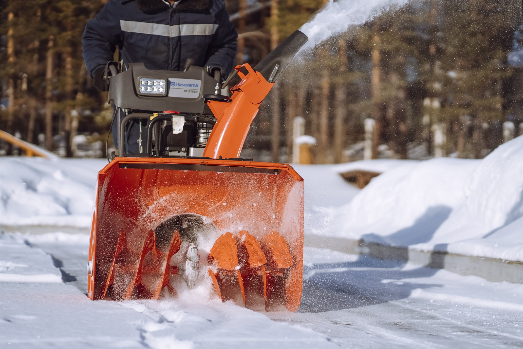 A person operates an orange snow blower, clearing fresh snow on a bright, sunny day, with a forested background.