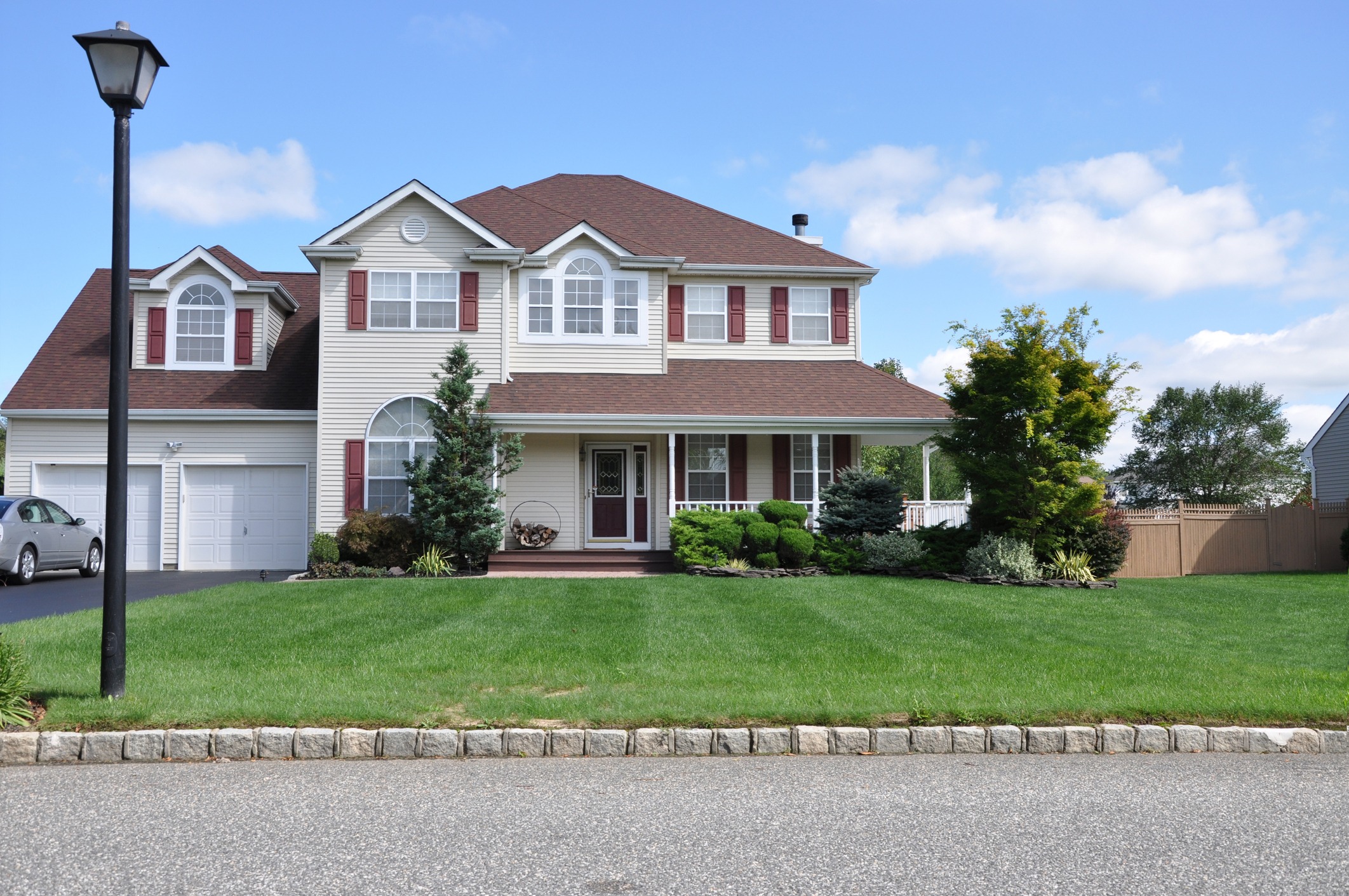 Suburban two-story house with brown roof, columns, and garage. Well-manicured lawn with a black lamp post and landscaped shrubs in foreground.