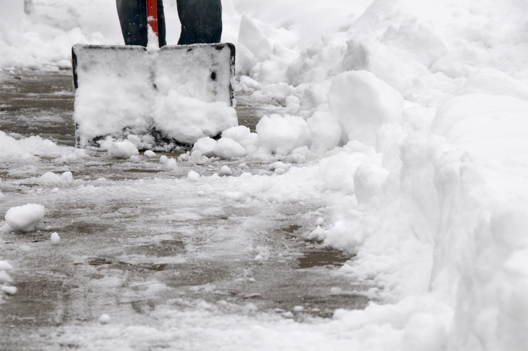 A person shovels snow from a sidewalk, creating a clear path amidst heavy snowfall, with large snow piles on either side.