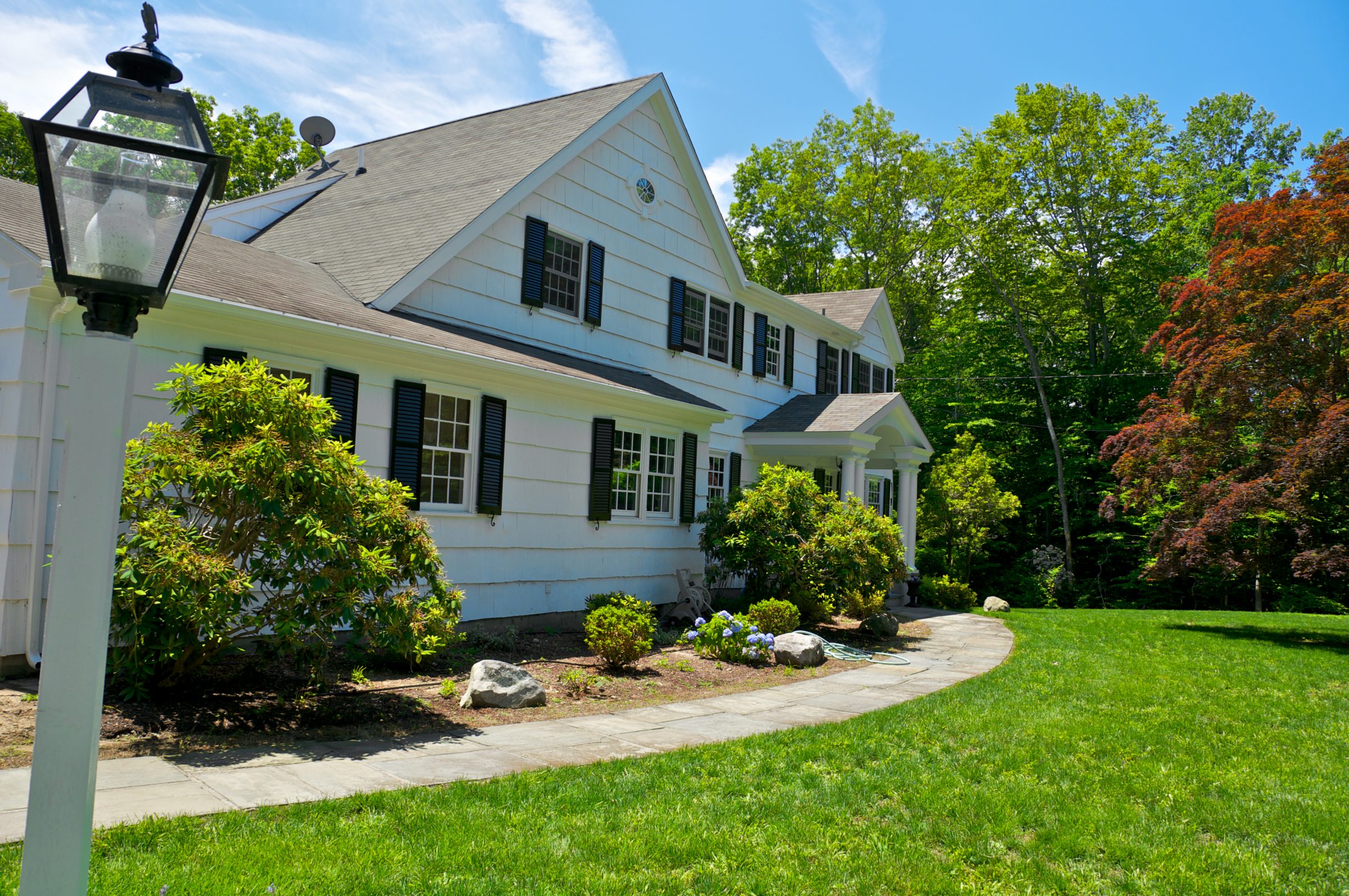A white house with black shutters, surrounded by lush greenery and a lamppost, sits under a clear blue sky.