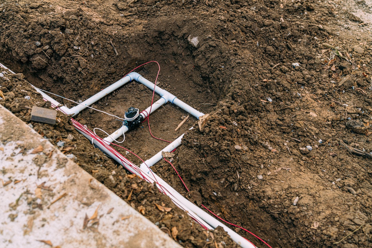 A trench with exposed white PVC pipes and red electrical wires, surrounded by soil, indicating ongoing construction or plumbing work.