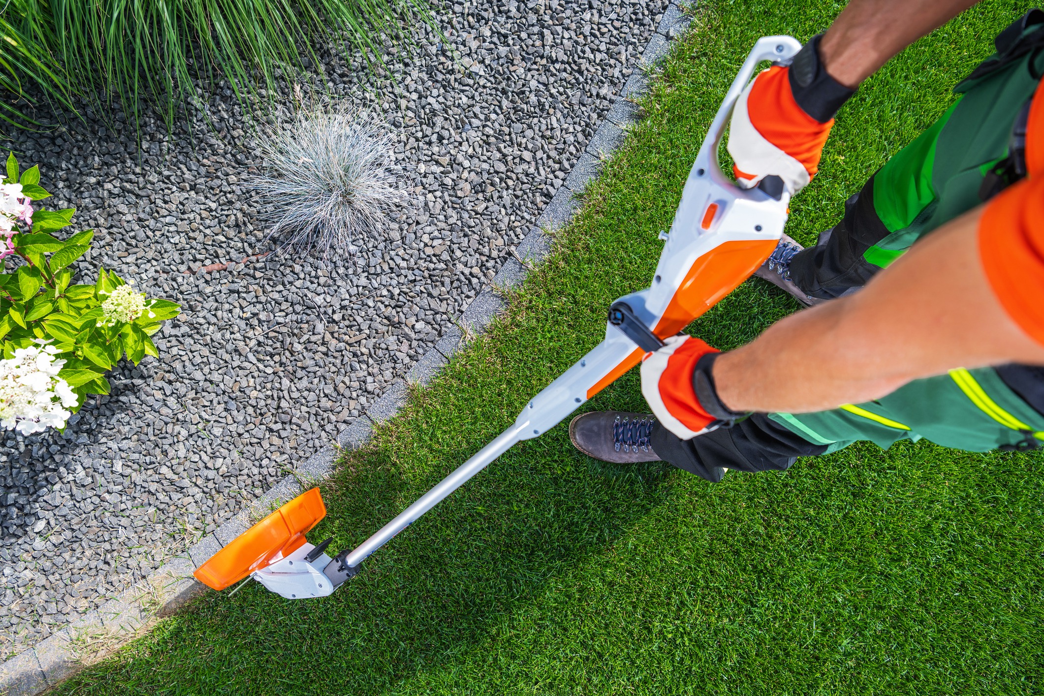 A person in gardening attire uses an orange trimmer to edge a lawn, surrounded by gravel and blooming plants.