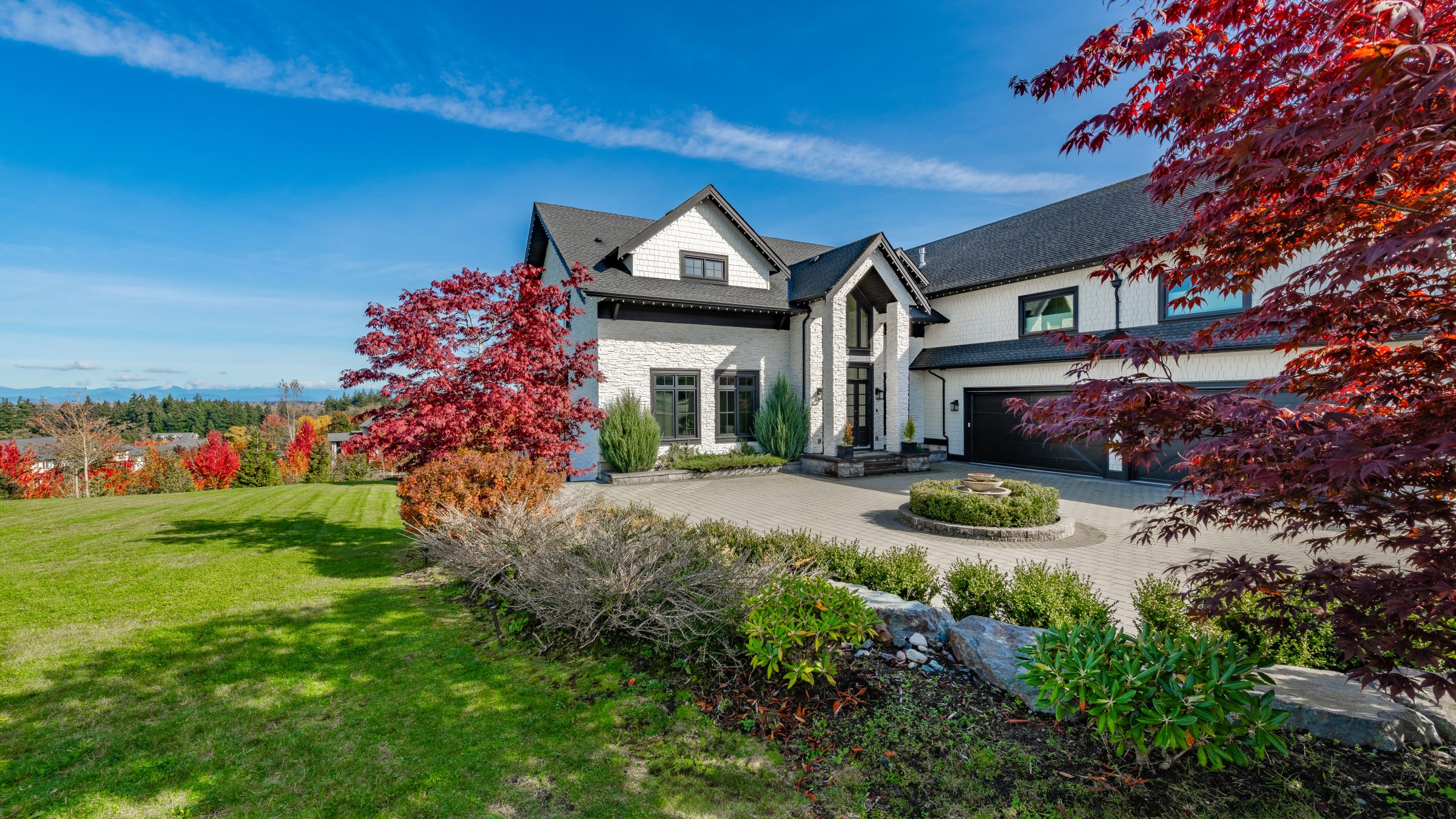 Modern house with white brick exterior, circular driveway, and landscaped garden. Surrounded by green lawn and red foliage under a bright blue sky.