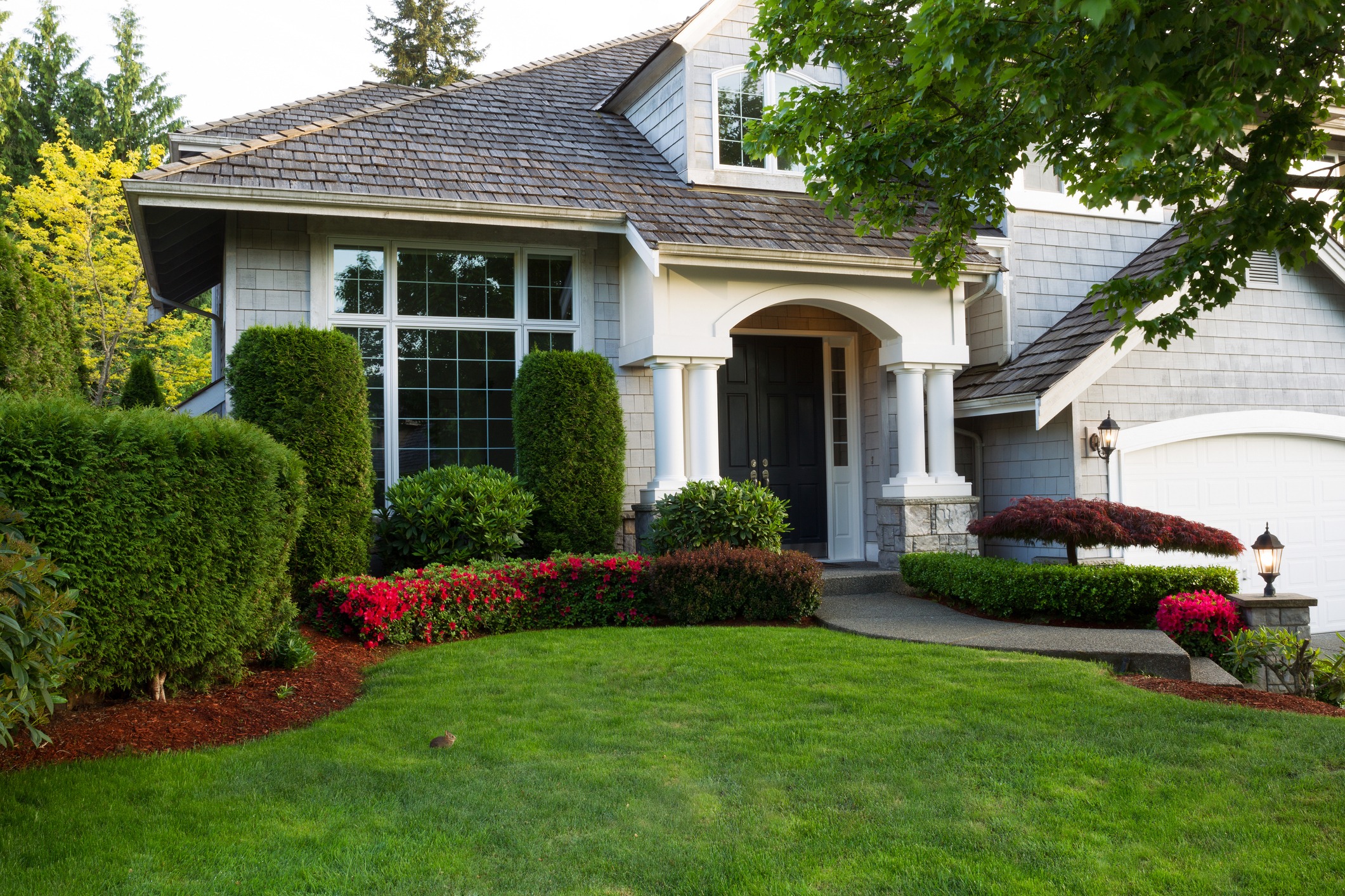 A charming suburban house with a manicured lawn, blooming flowers, and lush trees. Traditional architecture features arched entrance and white columns.