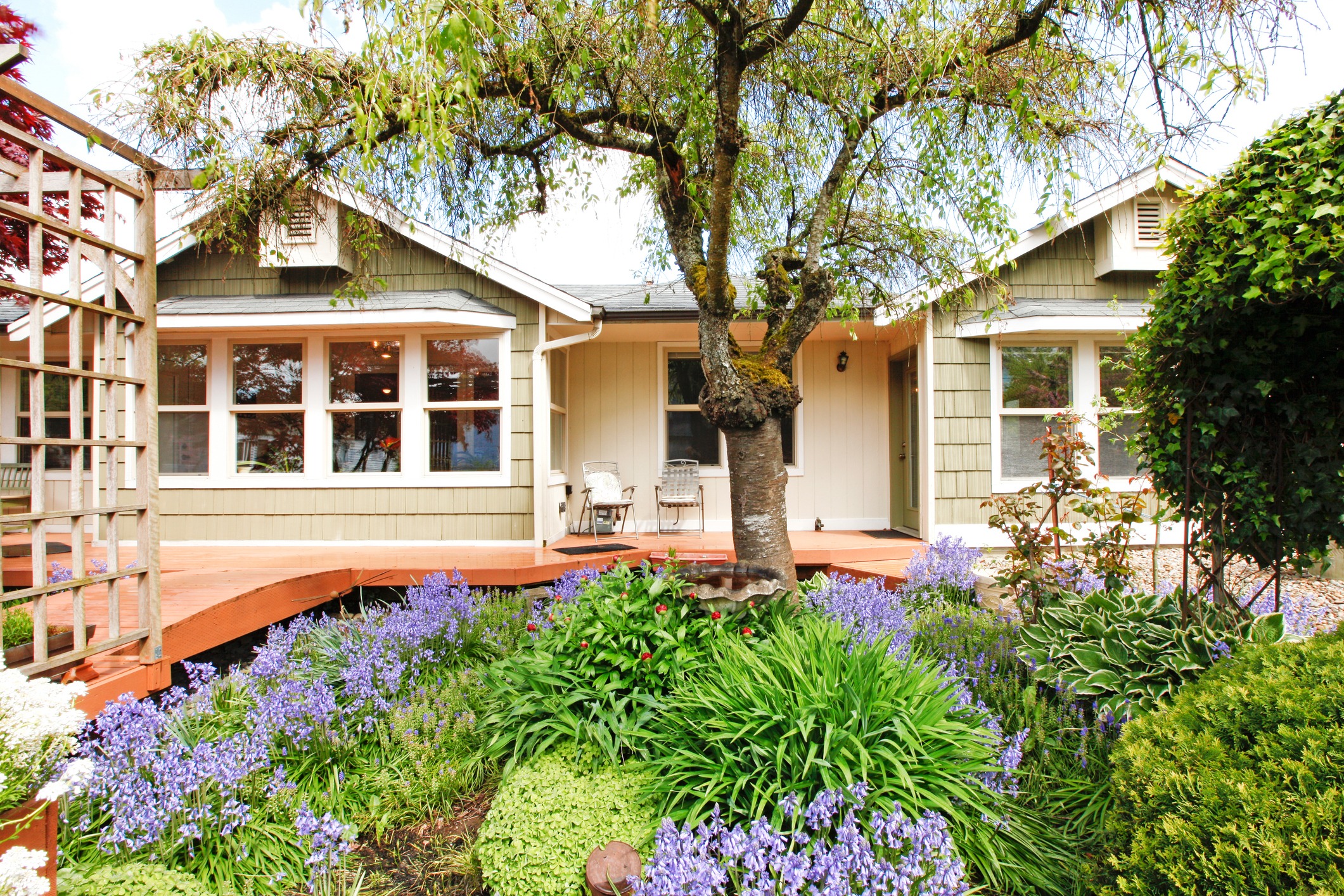 Clapboard siding house exterior. View of wooden deck and flower bed with blooming plants and trees