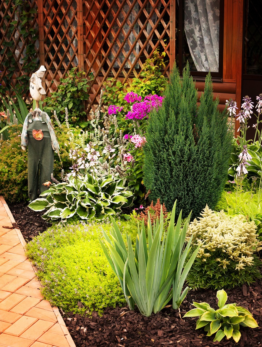 A lush garden with diverse plants, colorful flowers, a wooden lattice backdrop, and a whimsical scarecrow beside a brick pathway.
