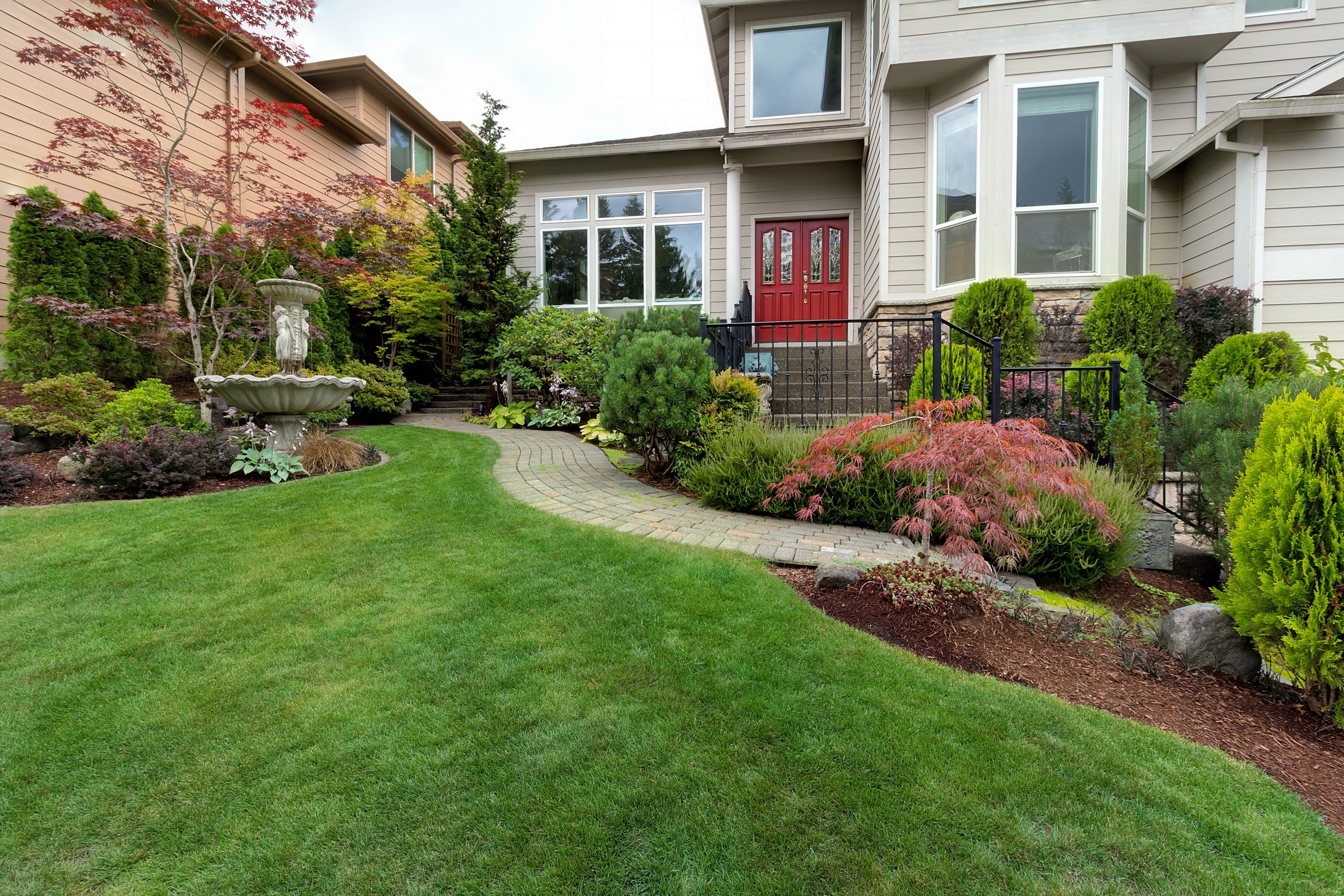 Well-maintained garden with a stone path, fountain, and colorful shrubs leads to a modern house with red doors and large windows.