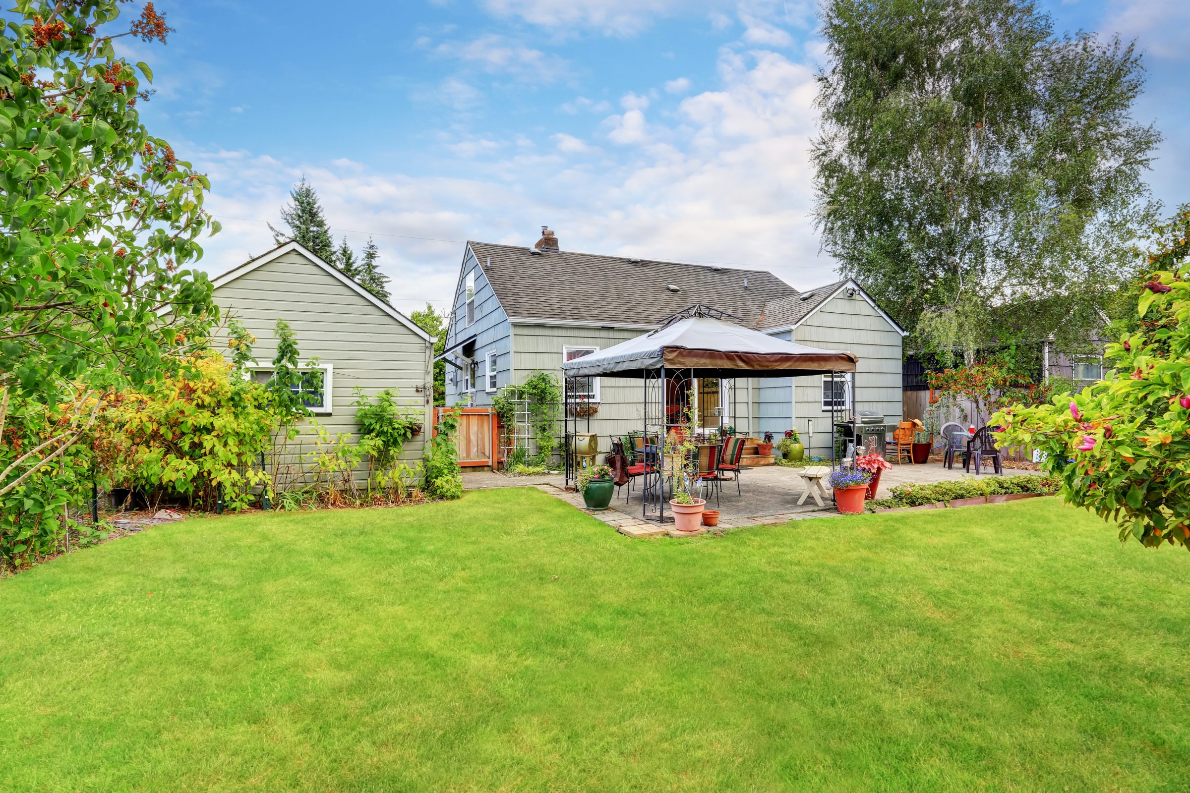 A cozy backyard with lush grass, trees, and patio furniture under a gazebo, adjacent to a charming two-story house with small garden.