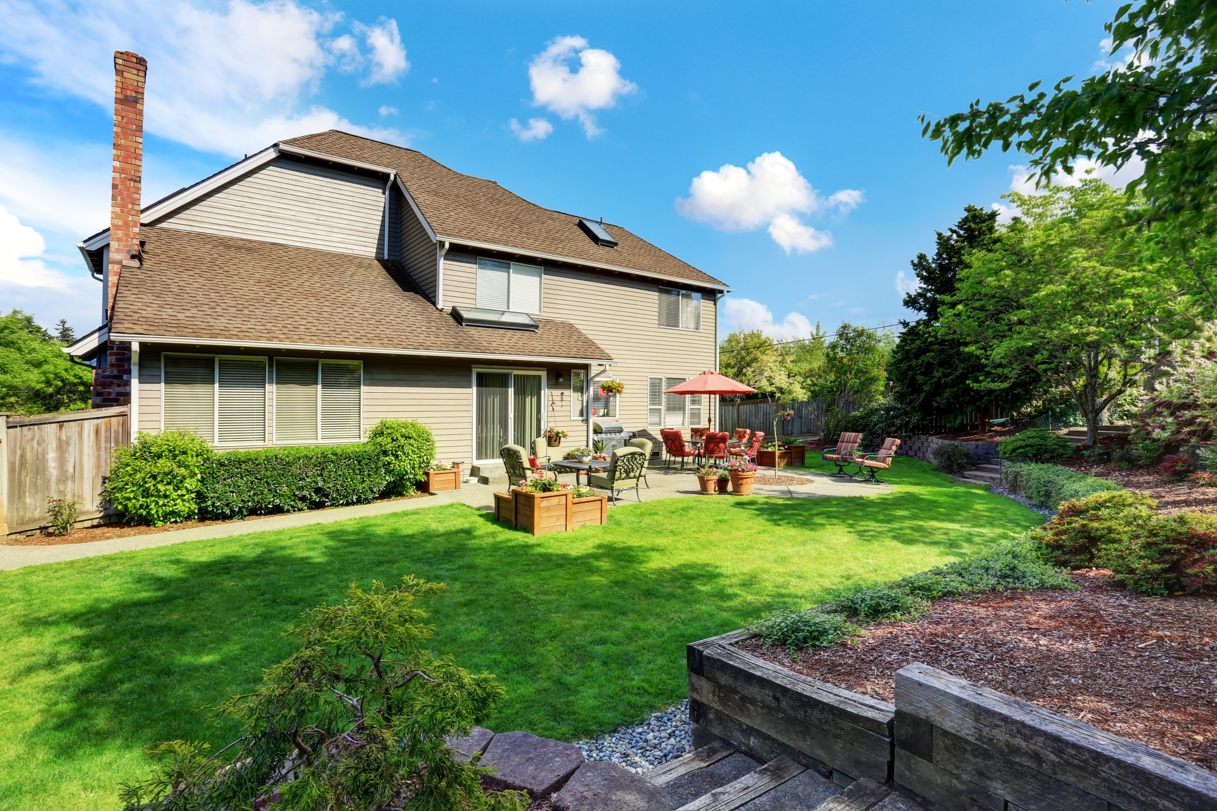 A suburban backyard with green lawn, patio furniture, red umbrella, potted plants, and a modern house under a sunny blue sky.