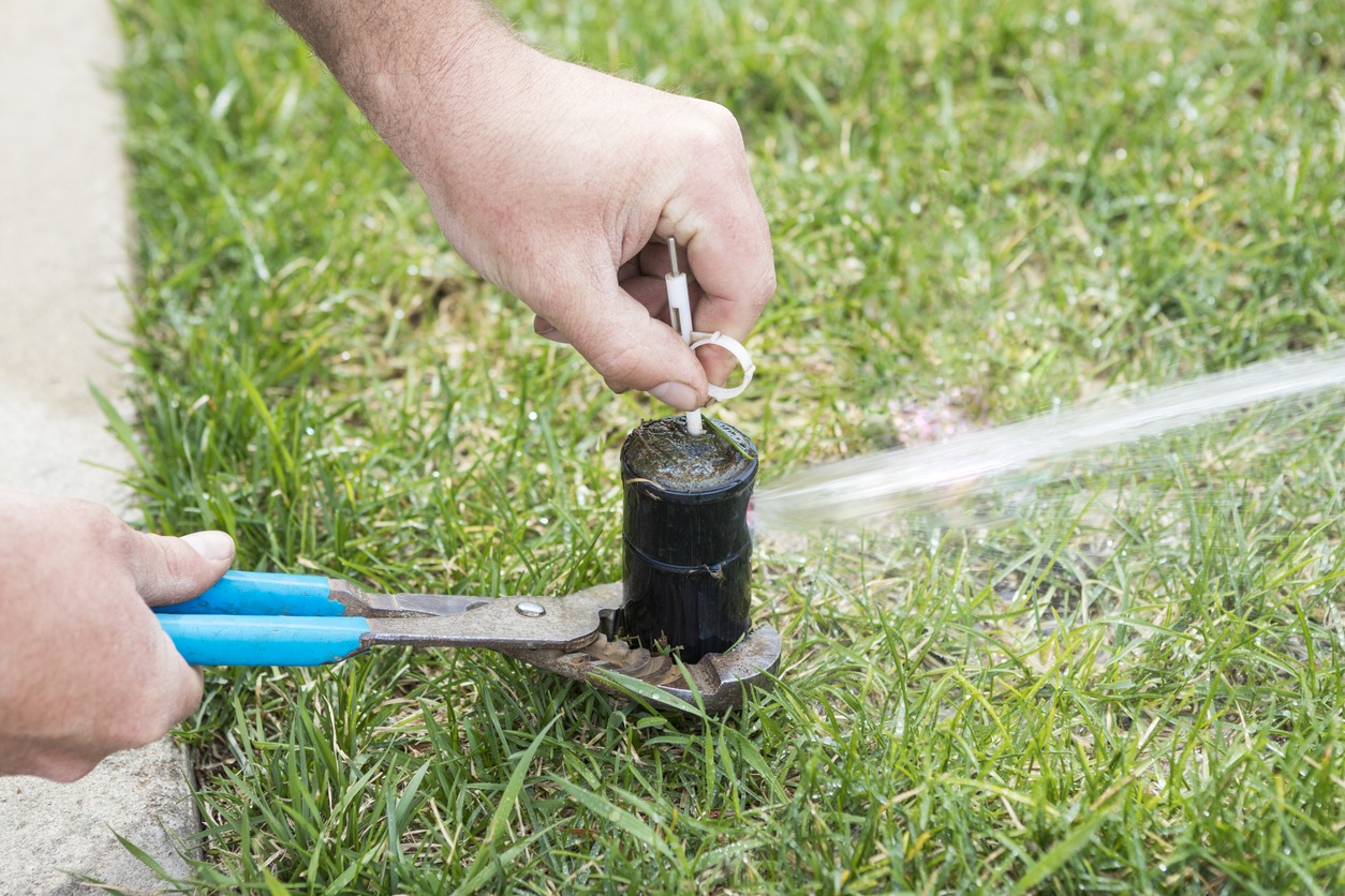 A person uses pliers to adjust a sprinkler head in a grassy area, with water spraying in a steady stream to the right.