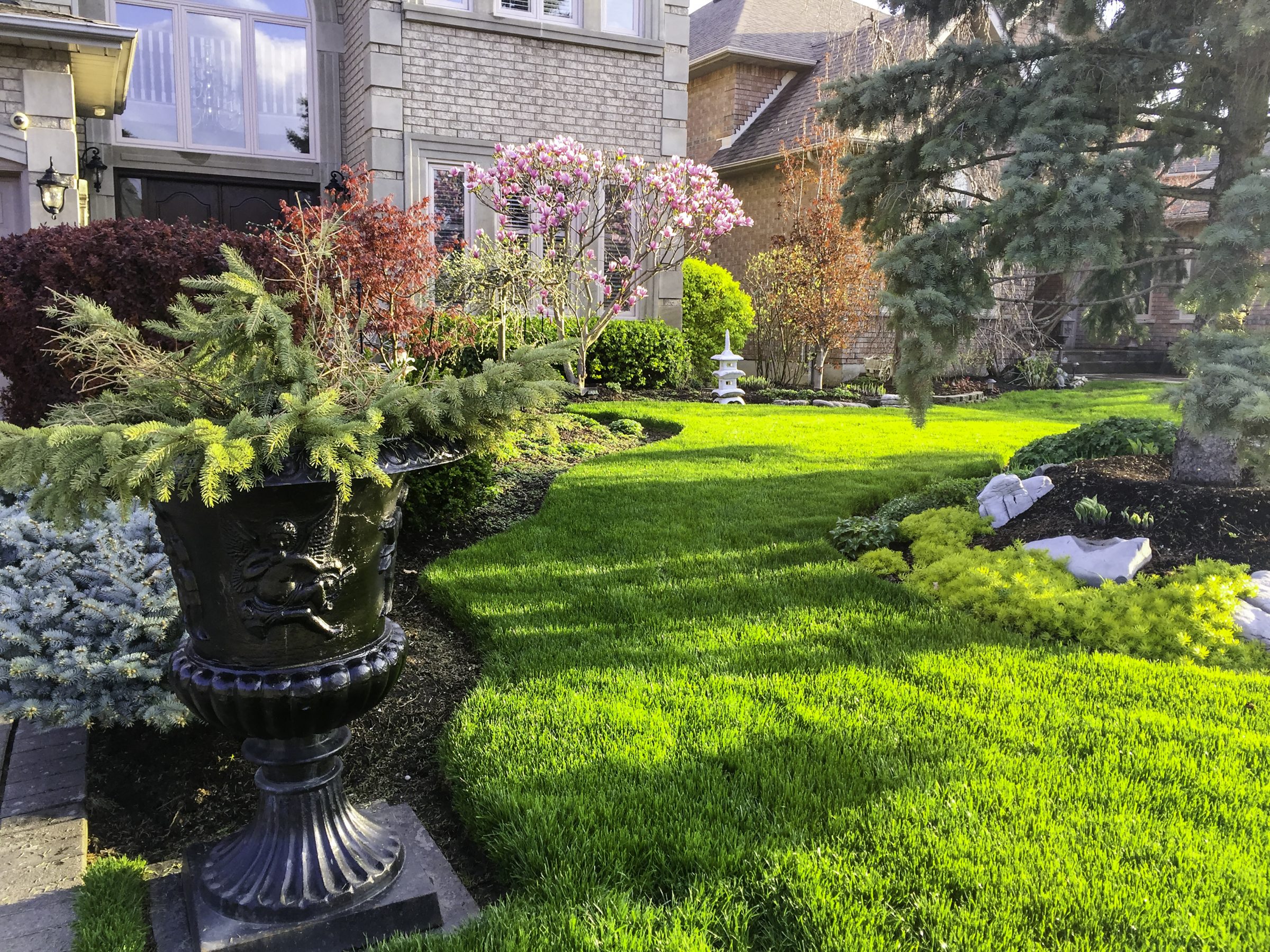 A well-manicured lawn with lush greenery surrounds a residential building. Decorative elements include a large planter and ornamental stones, creating an elegant garden.